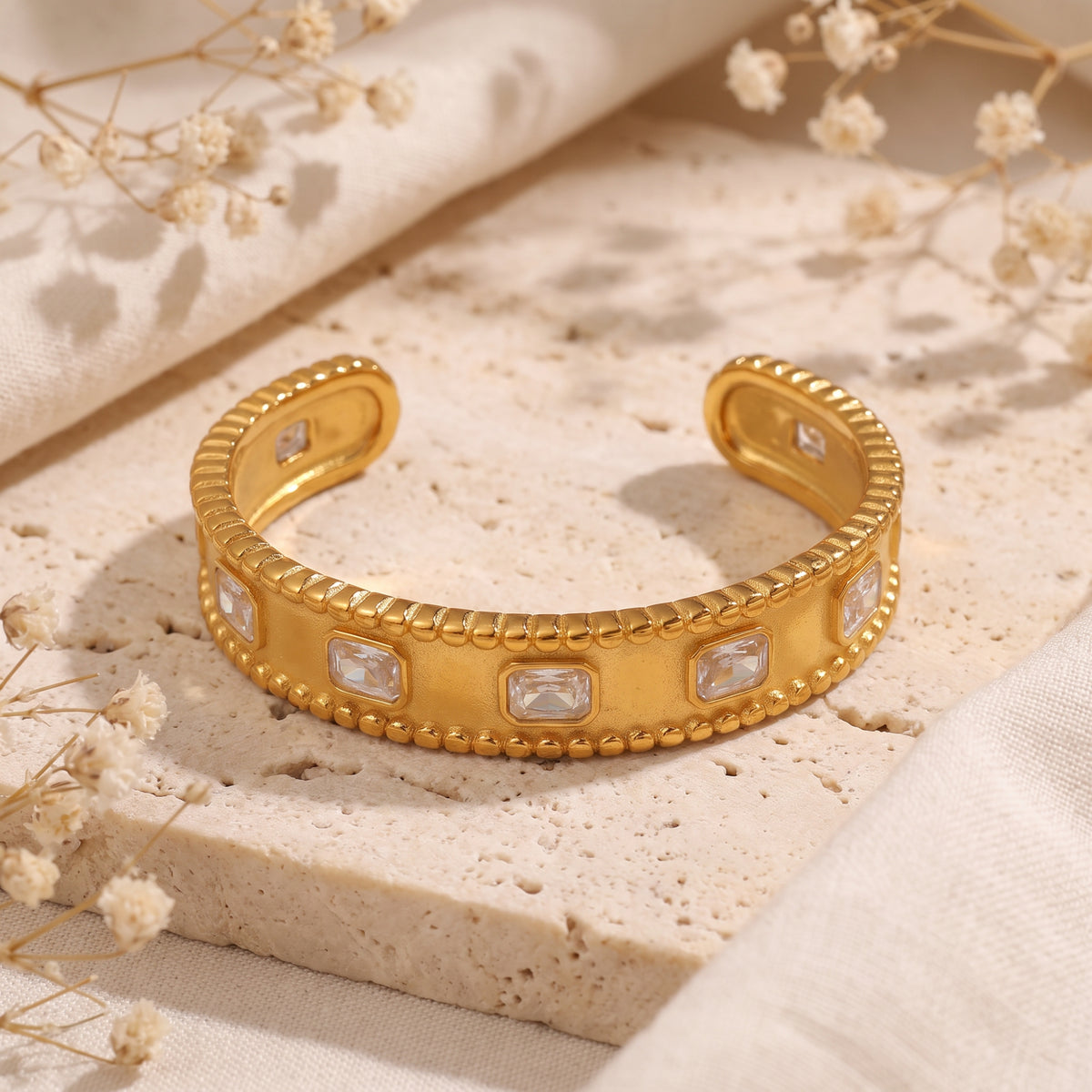 Open gold-tone cuff bracelet with beaded edges and clear rectangular stones displayed on a stone surface.