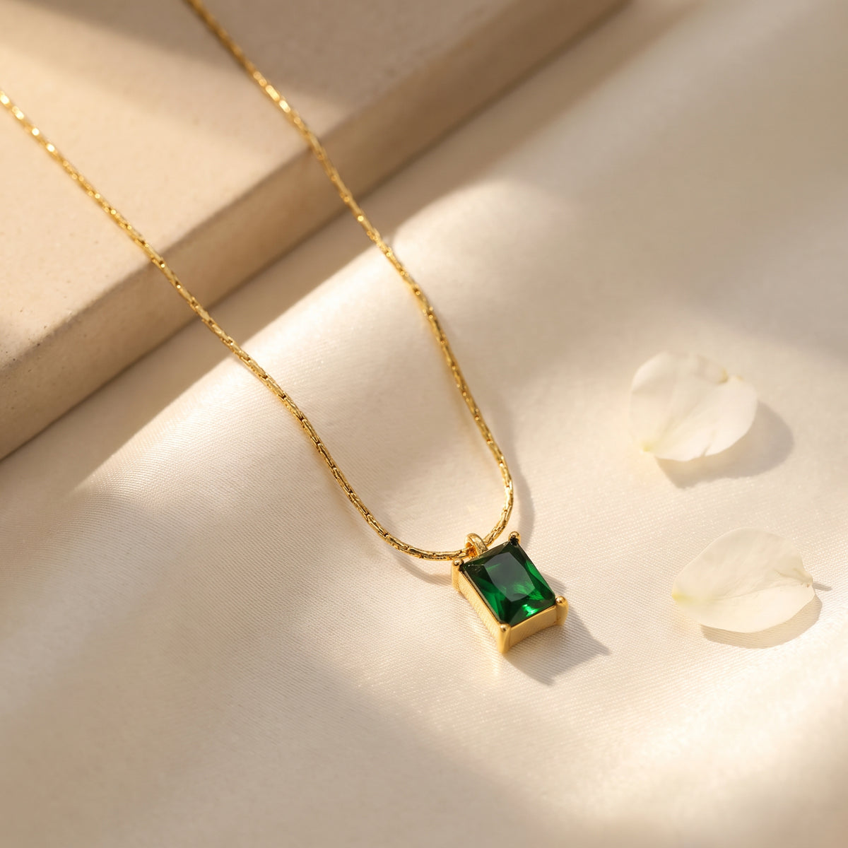 Angled necklace view on light fabric, showing the gold-tone chain and green rectangular pendant beside white petals.