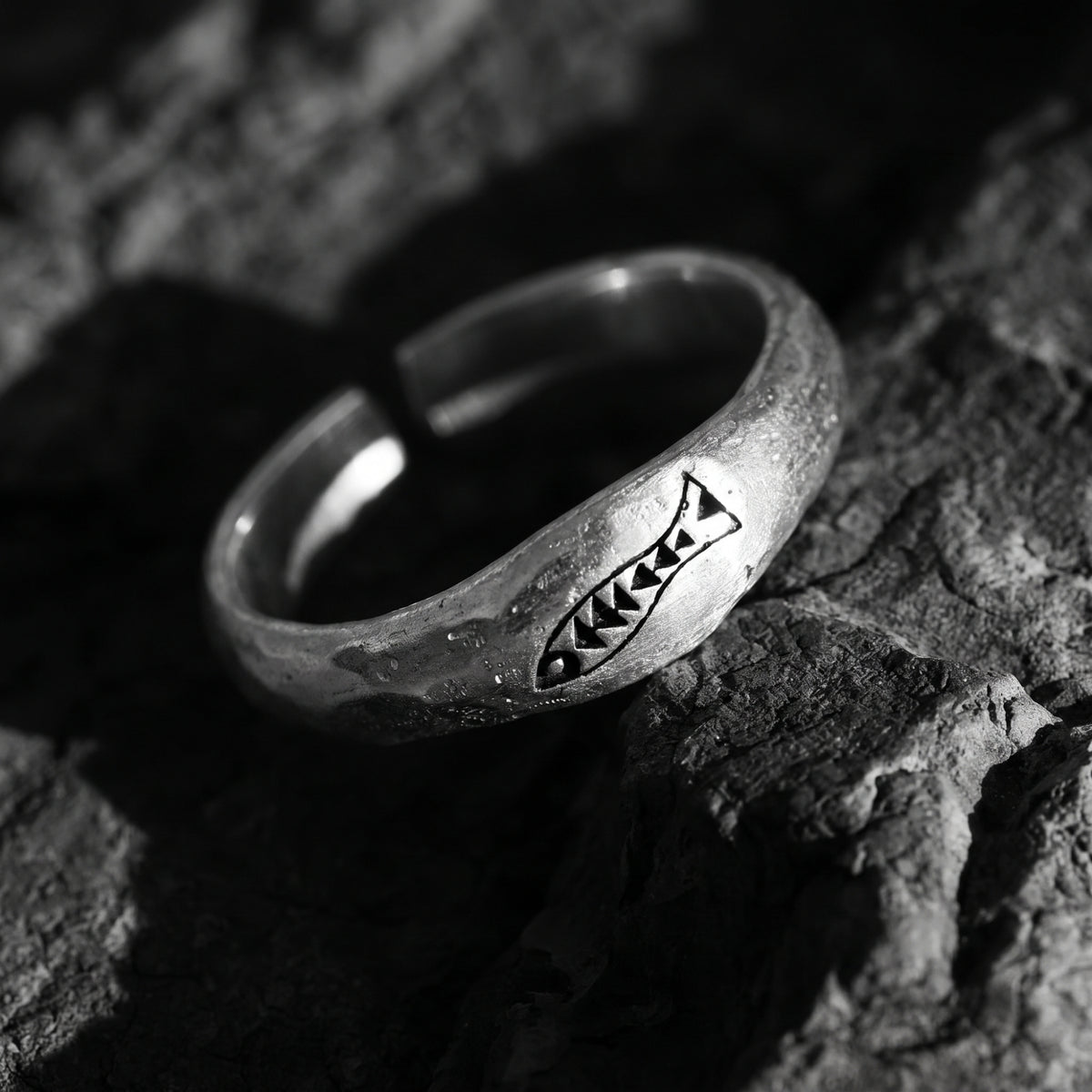 Angled black-and-white view of a silver carved fish ring showing its open back on rough stone.