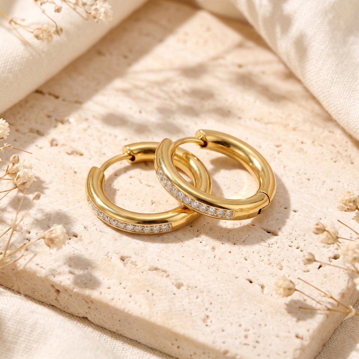 Pair of gold-tone pavé huggie hoop earrings overlapping on a beige textured stone surface.