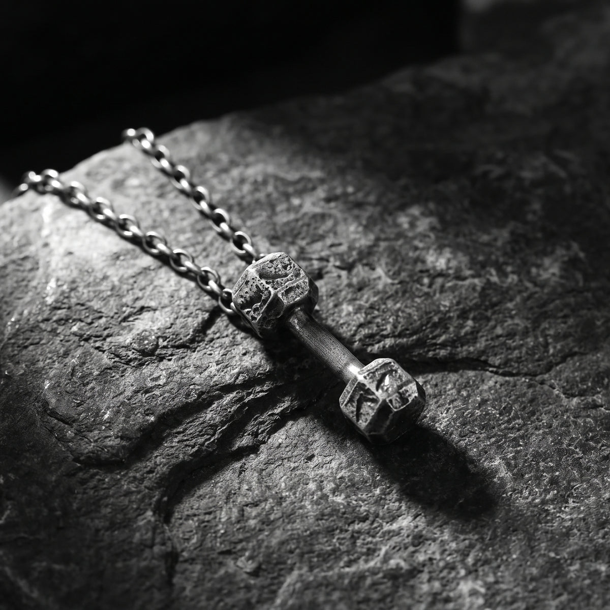 Fine silver dumbbell pendant and chain resting diagonally on a dark textured rock surface.