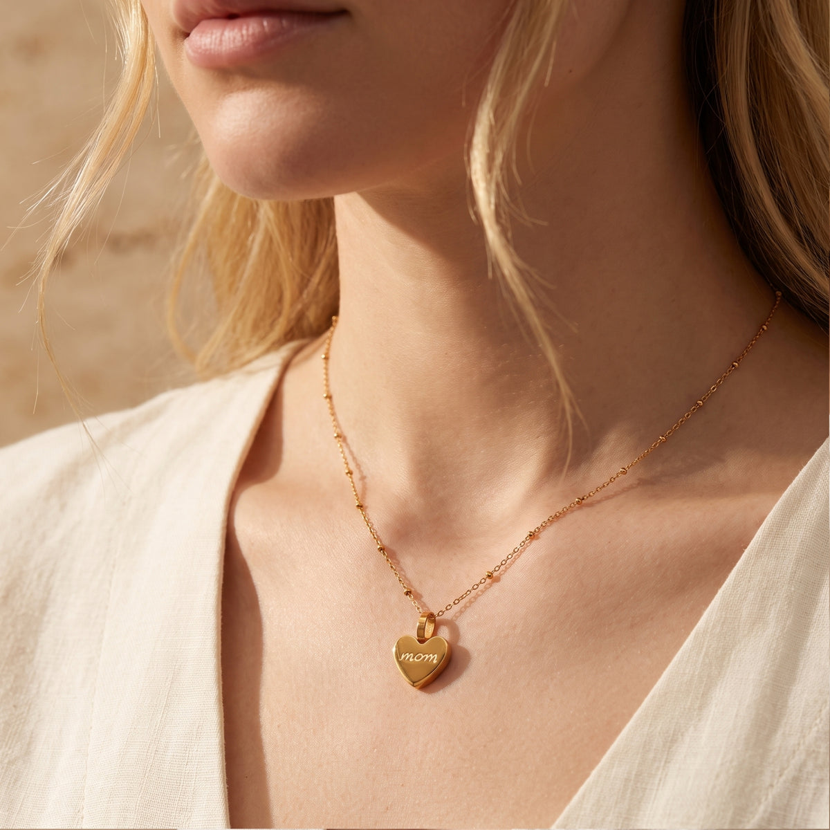 Model wearing a gold-tone heart pendant engraved with 'mom' on a ball-station chain with a light V-neck top.