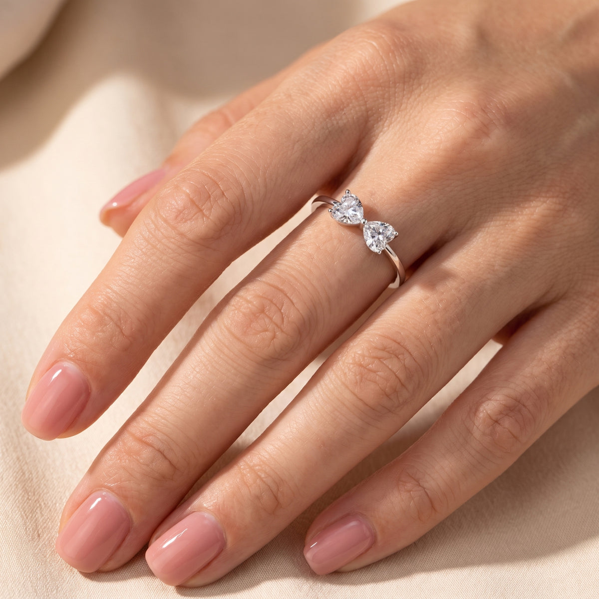 Full hand view displaying the silver bow ring with twin heart-shaped clear stones against silk.