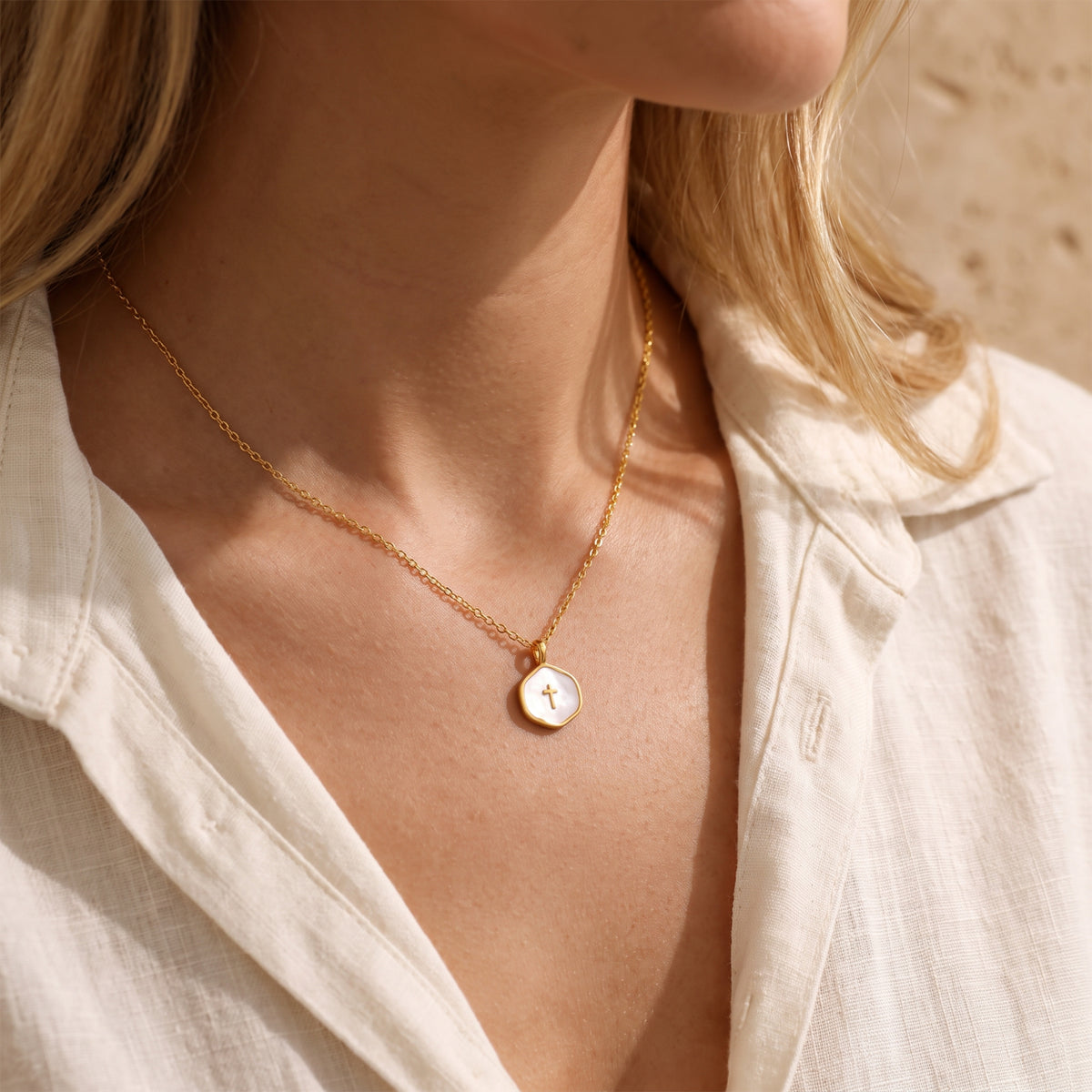 Model wearing a gold-tone necklace with a white enamel cross pendant over an open white linen shirt.