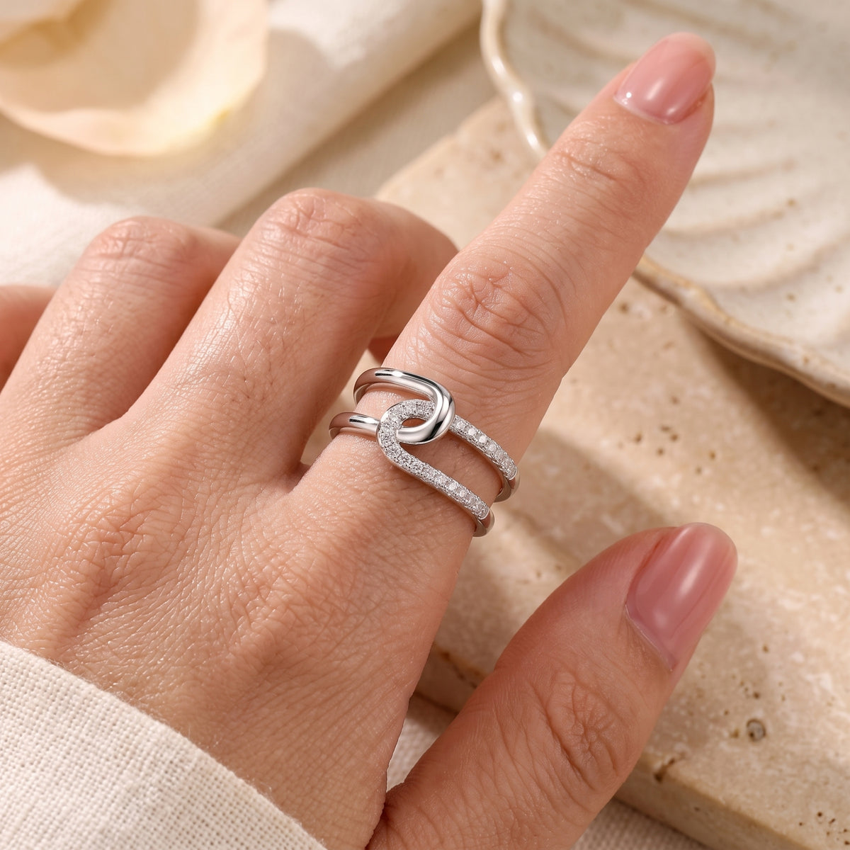 Close-up of the silver crossover knot ring with cubic zirconia worn on a finger against beige stone.