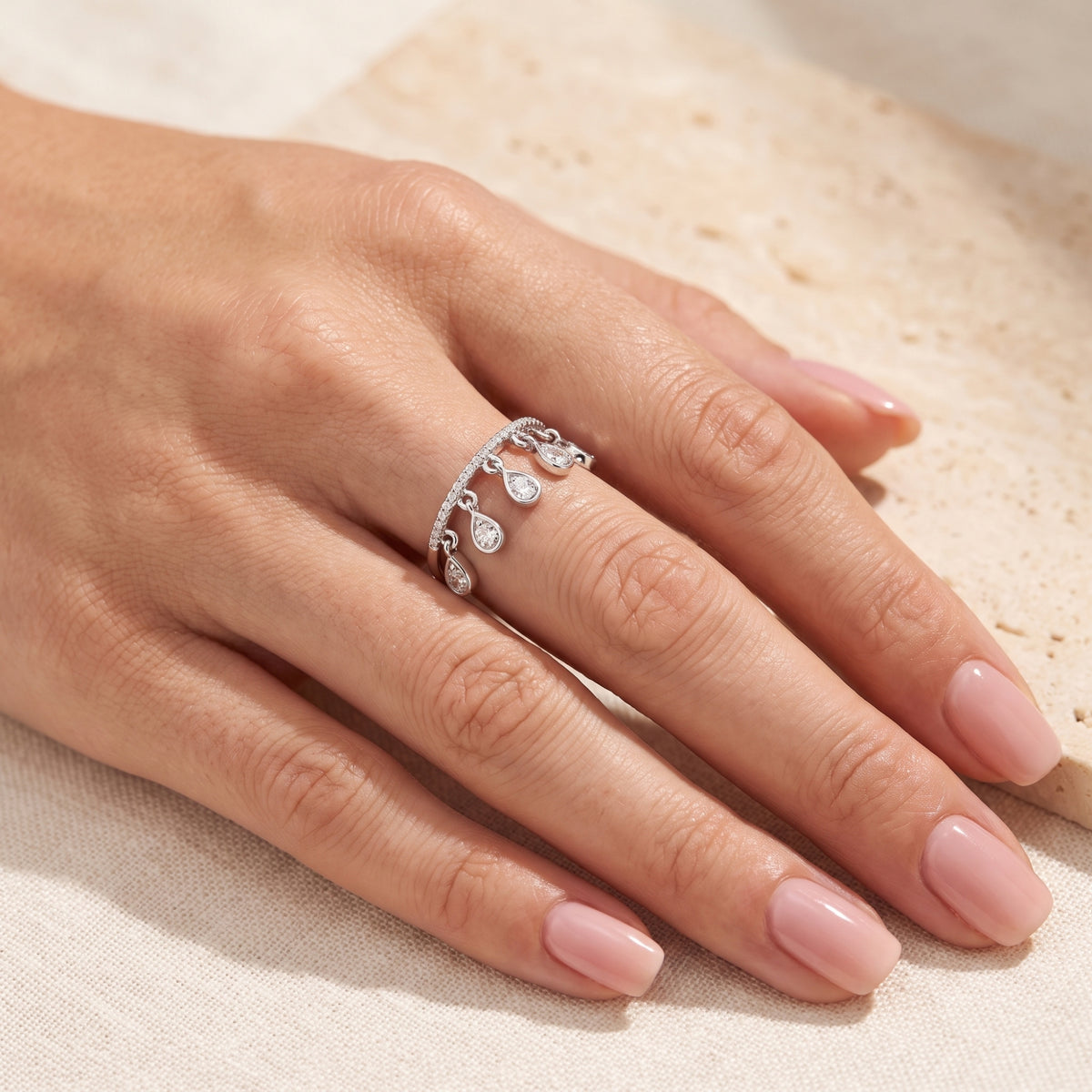 Close-up of a silver cascading teardrop charm ring worn on a hand over a cream textured surface.
