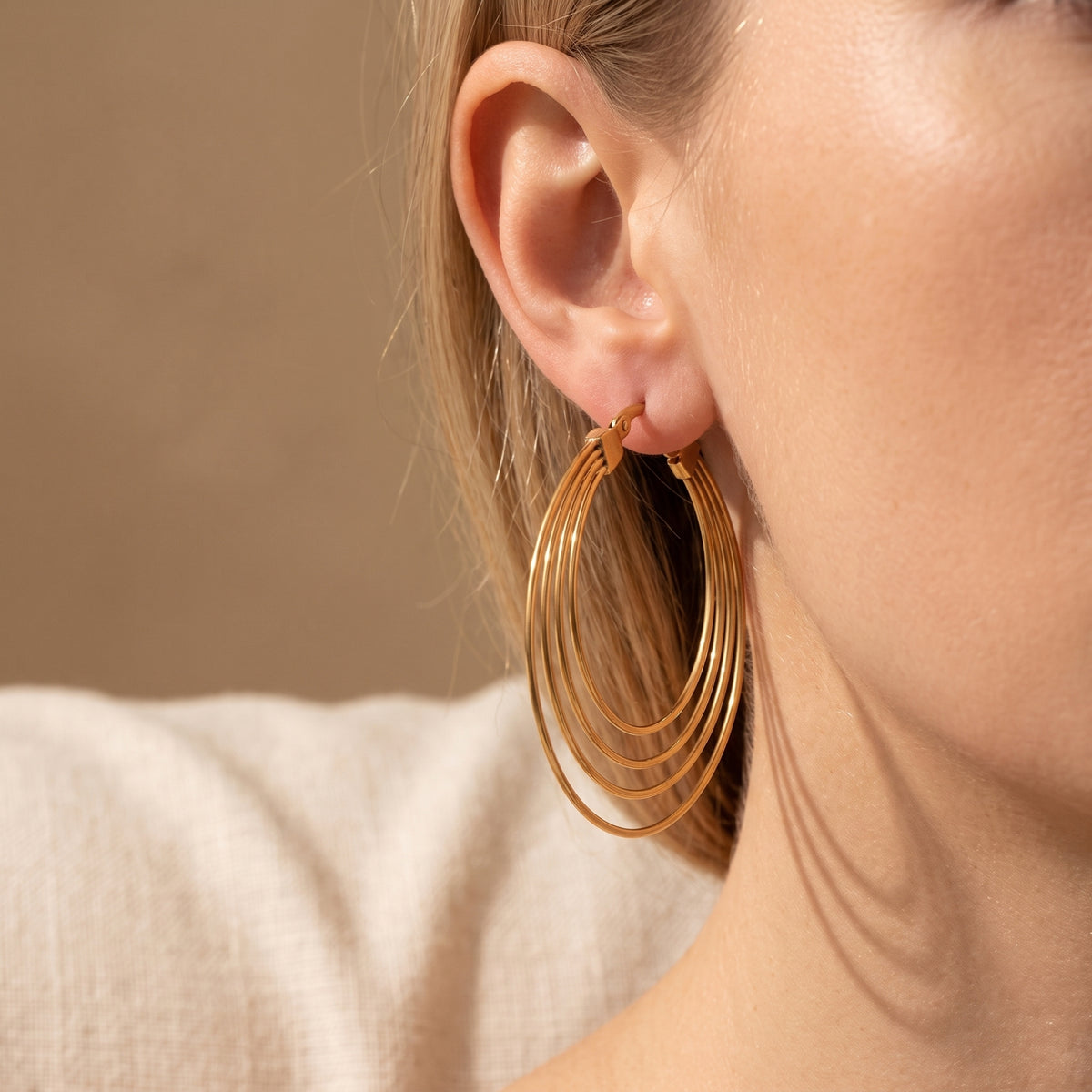 Side-profile close-up of a model wearing a gold-tone four-ring hoop earring with a light garment visible.