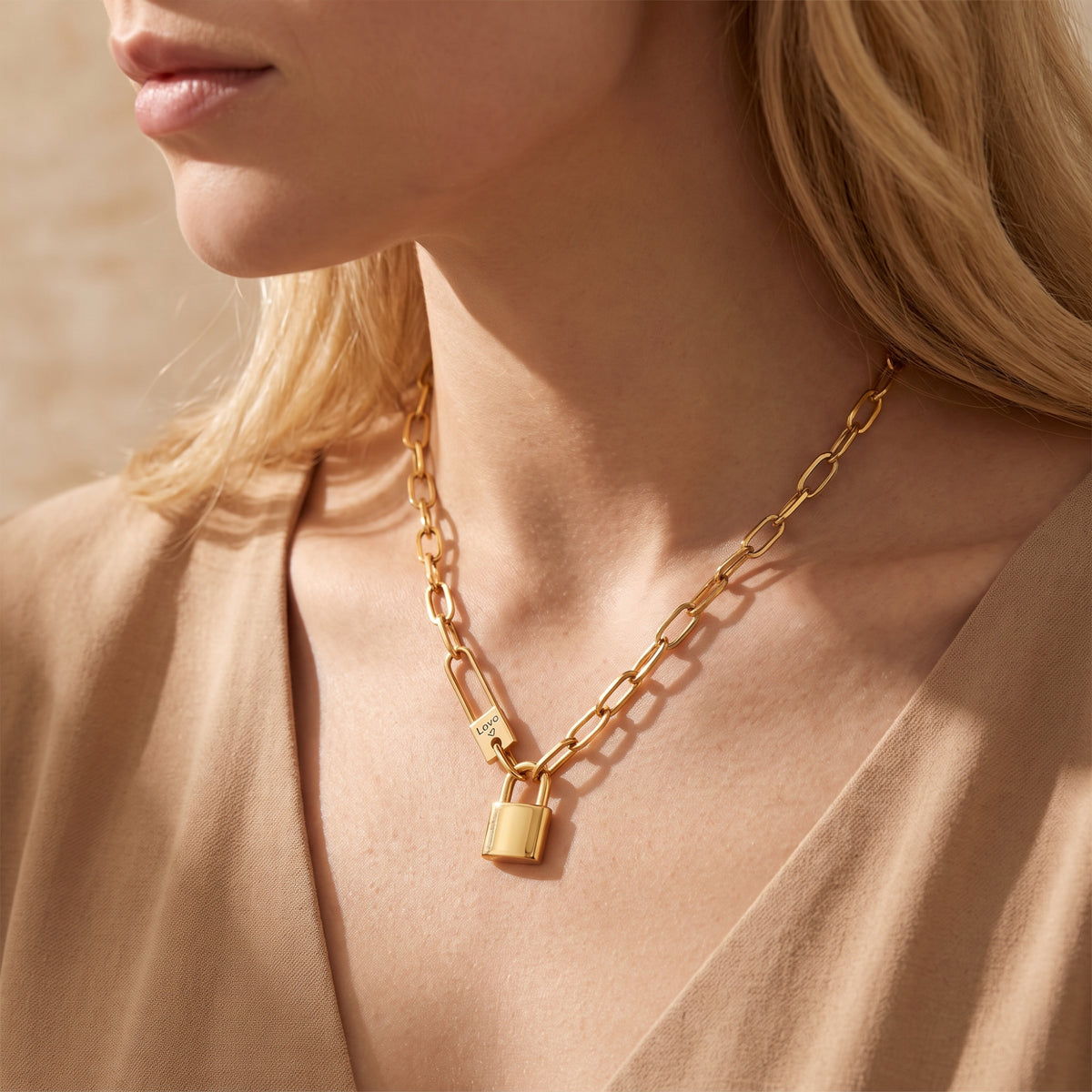 Close-up of a model wearing a gold-tone paperclip necklace with a padlock pendant over a tan open neckline.