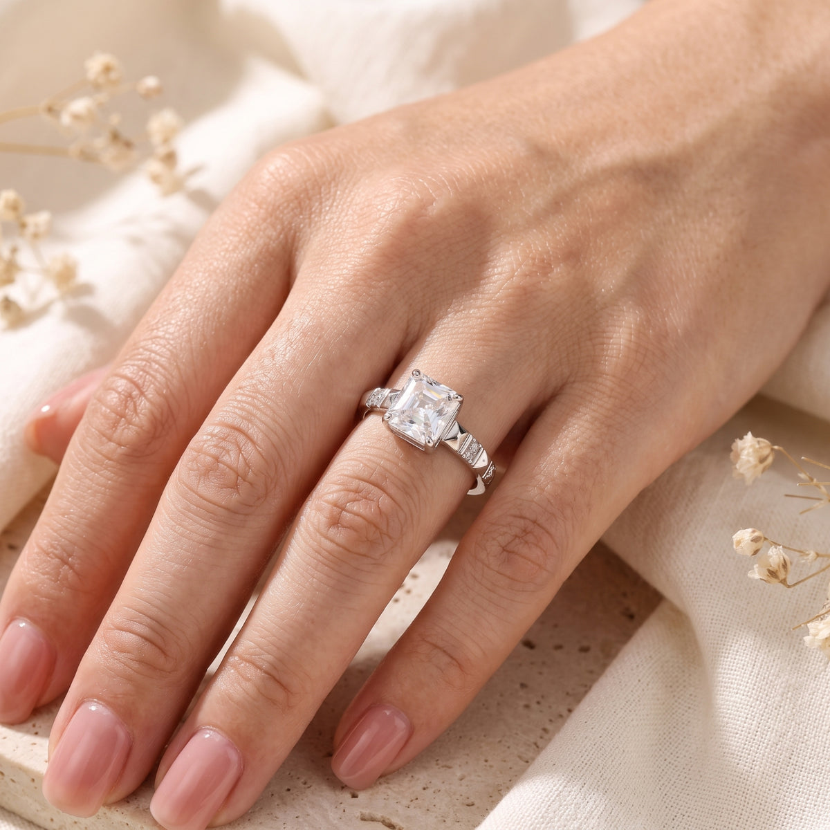 Angled hand view of the silver ring with a square radiant-cut stone worn against neutral stone and fabric.