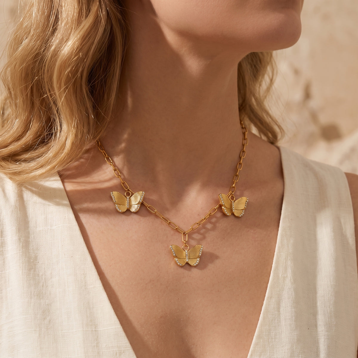 Model wearing the gold-tone three-butterfly necklace over a white open-neck garment, with charms across the chest.