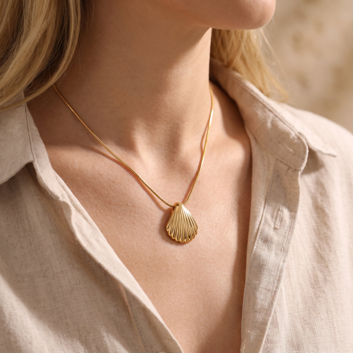 Close-up of the gold scallop shell pendant necklace worn with an open beige linen shirt.