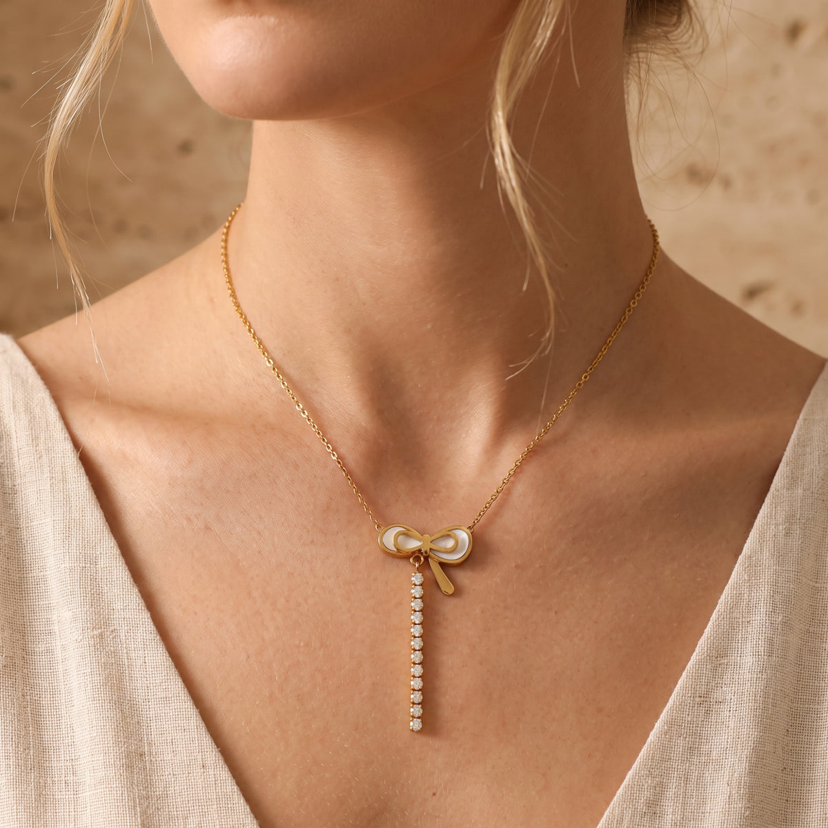Front view of the gold-tone bow necklace with pearlescent pendant and dangling clear stone drop worn over a beige top.