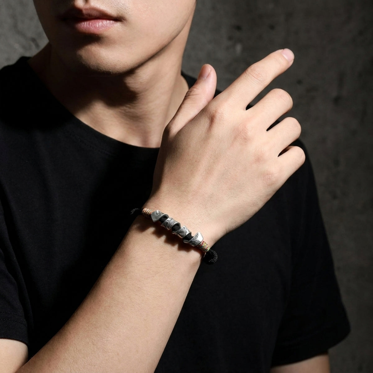 Person wearing a black cord bracelet with multiple engraved silver sections and multicolored thread accents on the wrist.