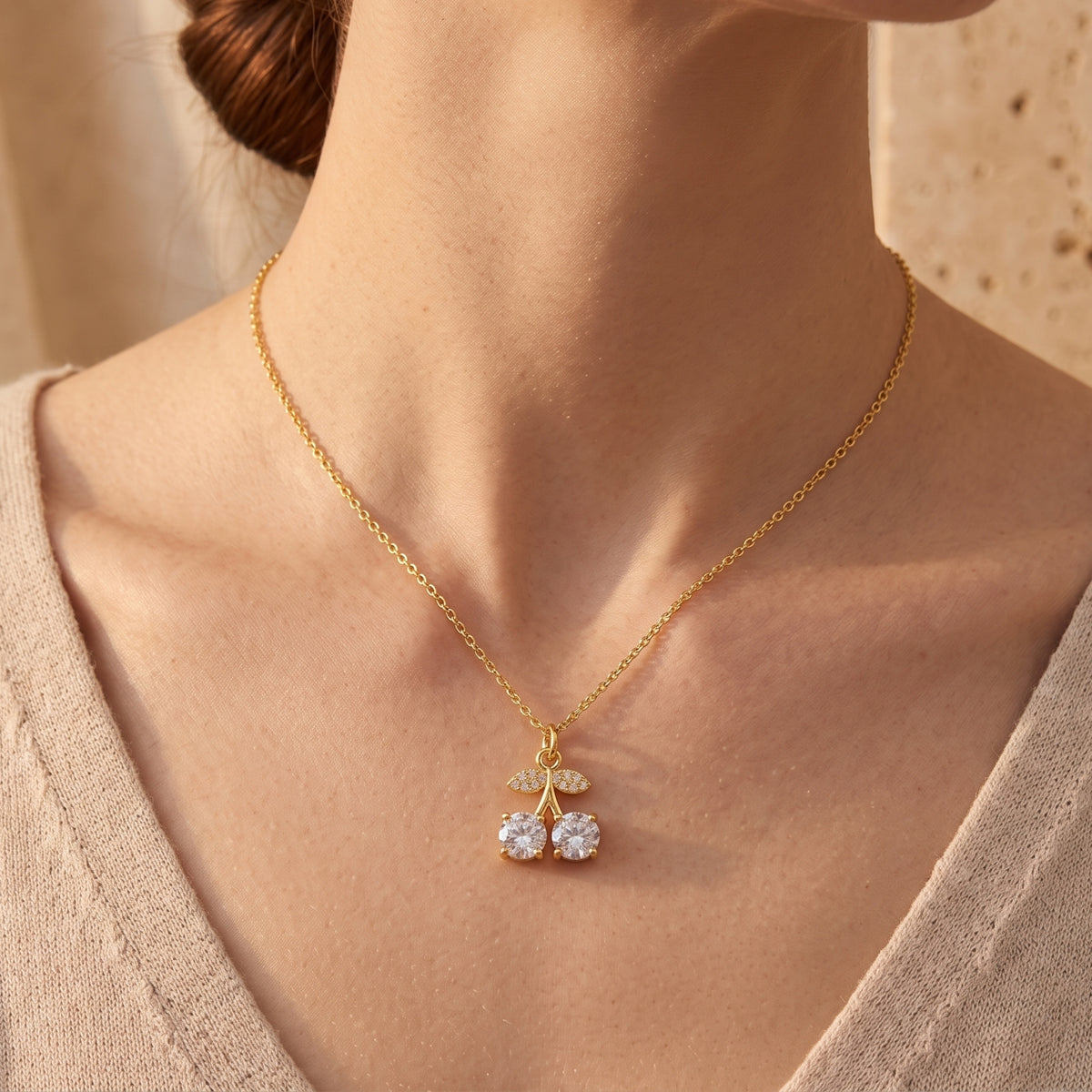 Gold-tone cherry pendant necklace shown on the back of a model's neck above a beige knit top.