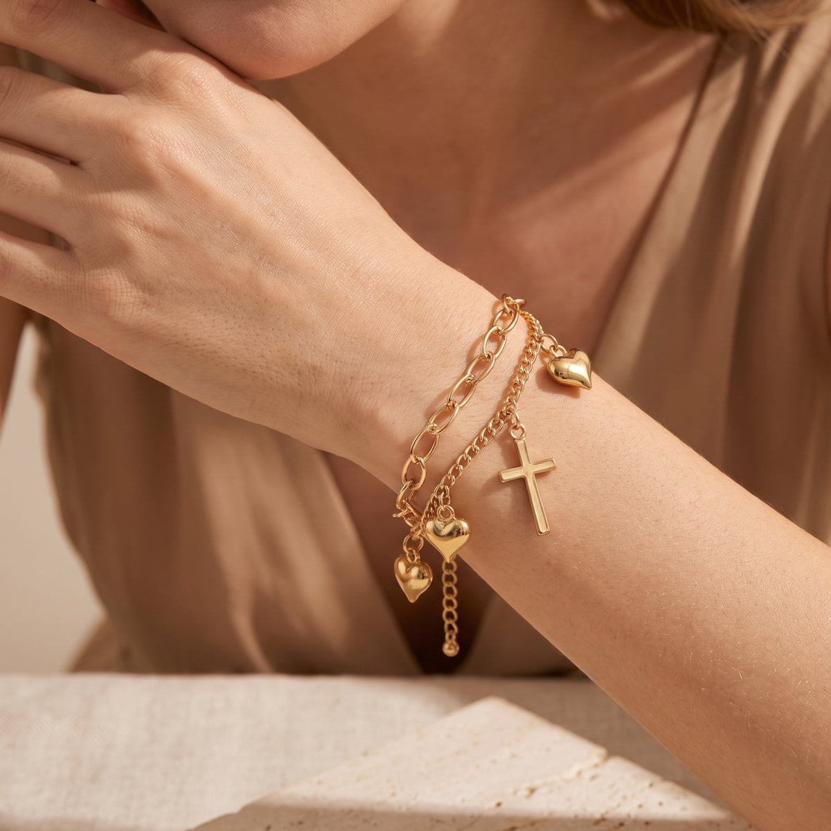 Model wearing a layered gold-tone charm bracelet with heart and cross charms against a beige outfit.