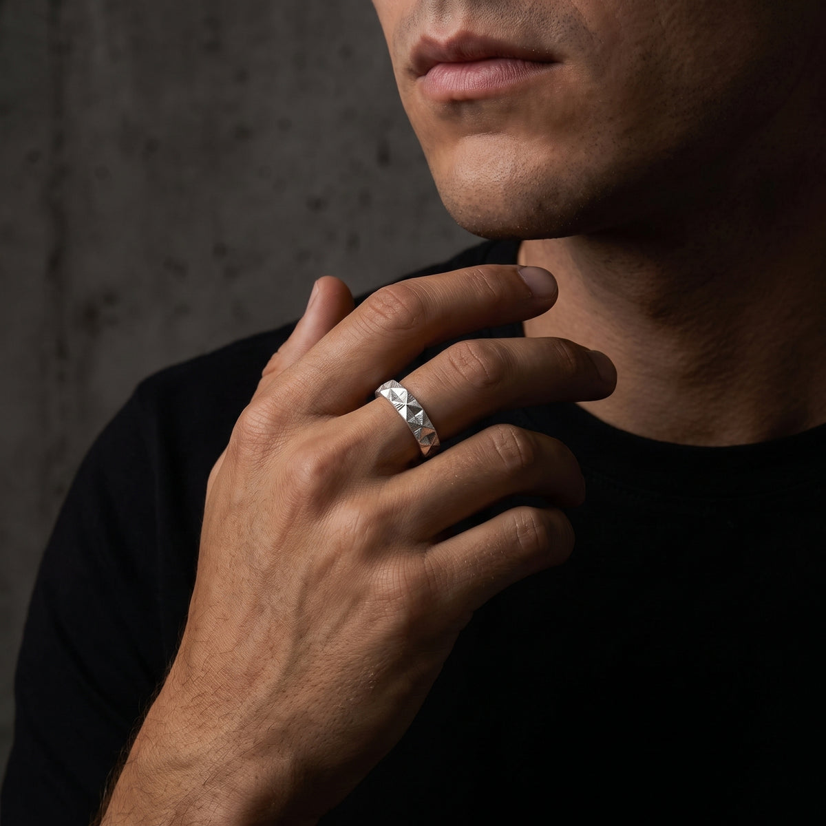 Man wearing the faceted silver pyramid ring on his middle finger near his neck against a dark concrete wall.