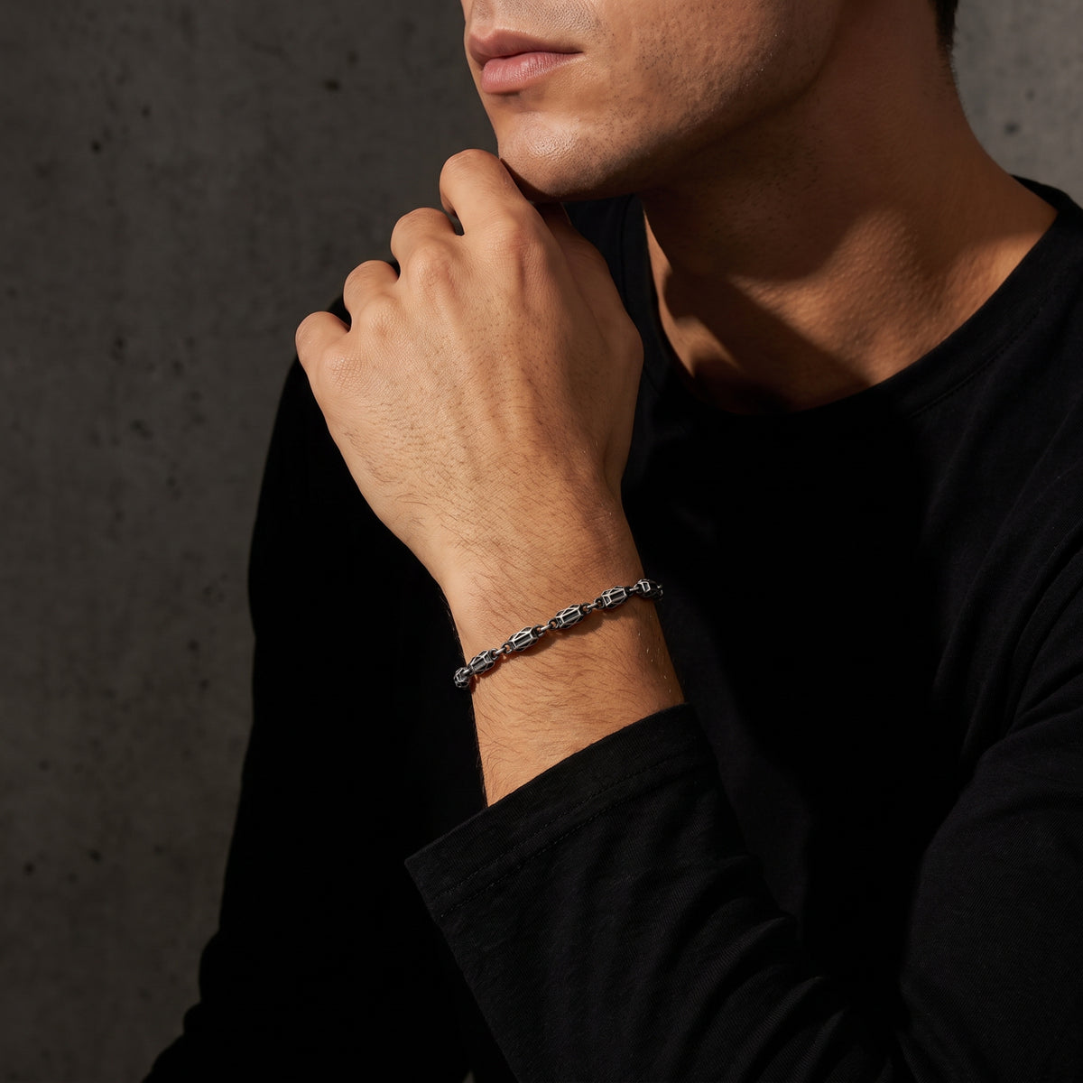 Man resting his hand near his chin, showing the silver geometric link bracelet over a black sleeve.