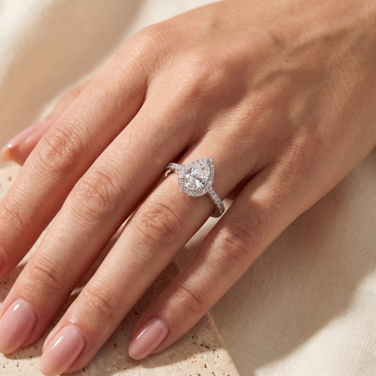 Top-down view of a hand wearing a silver pear-cut halo ring on the ring finger over a beige stone surface.