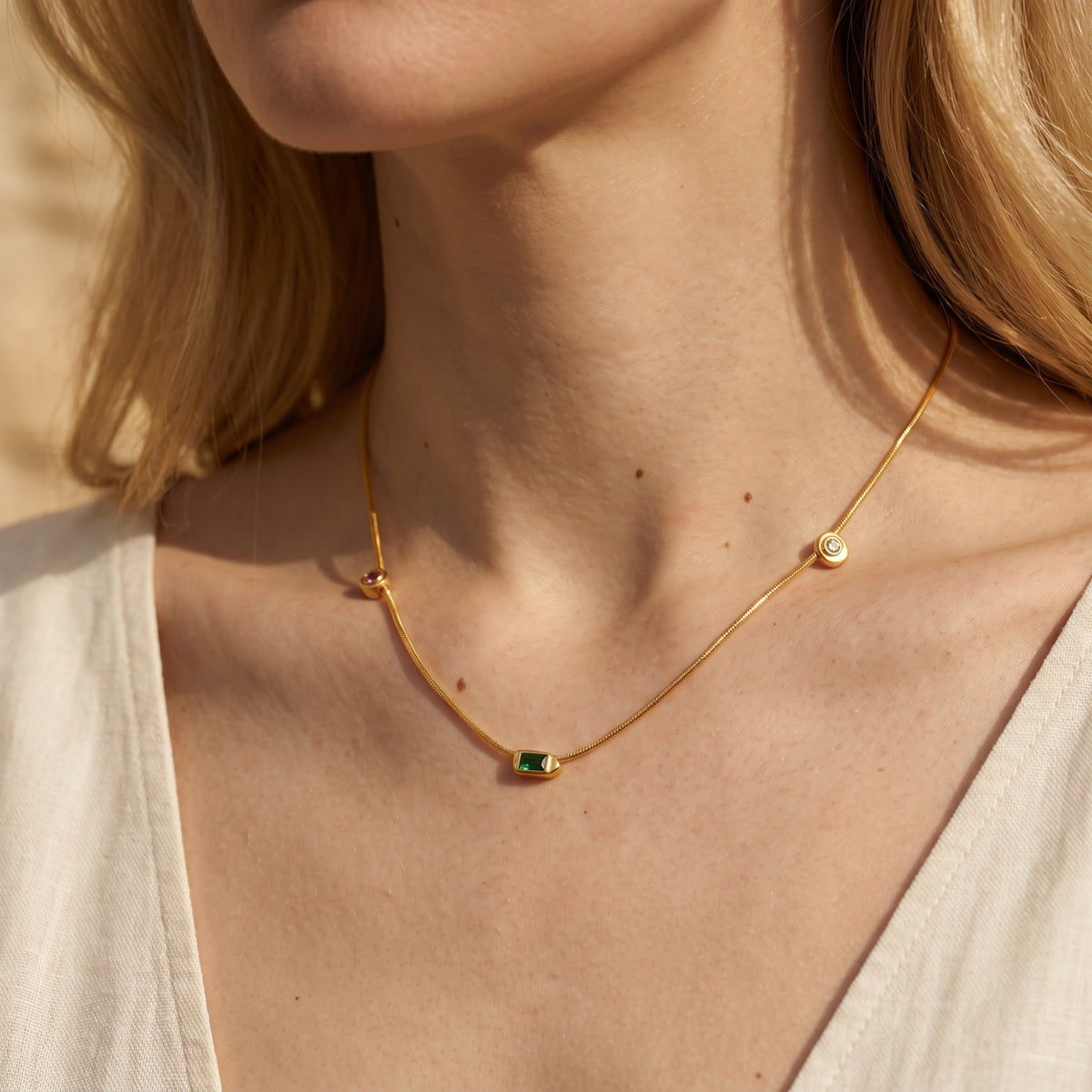 Angled on-model view of the gold station necklace with a green center stone and beige textured V-neck top.