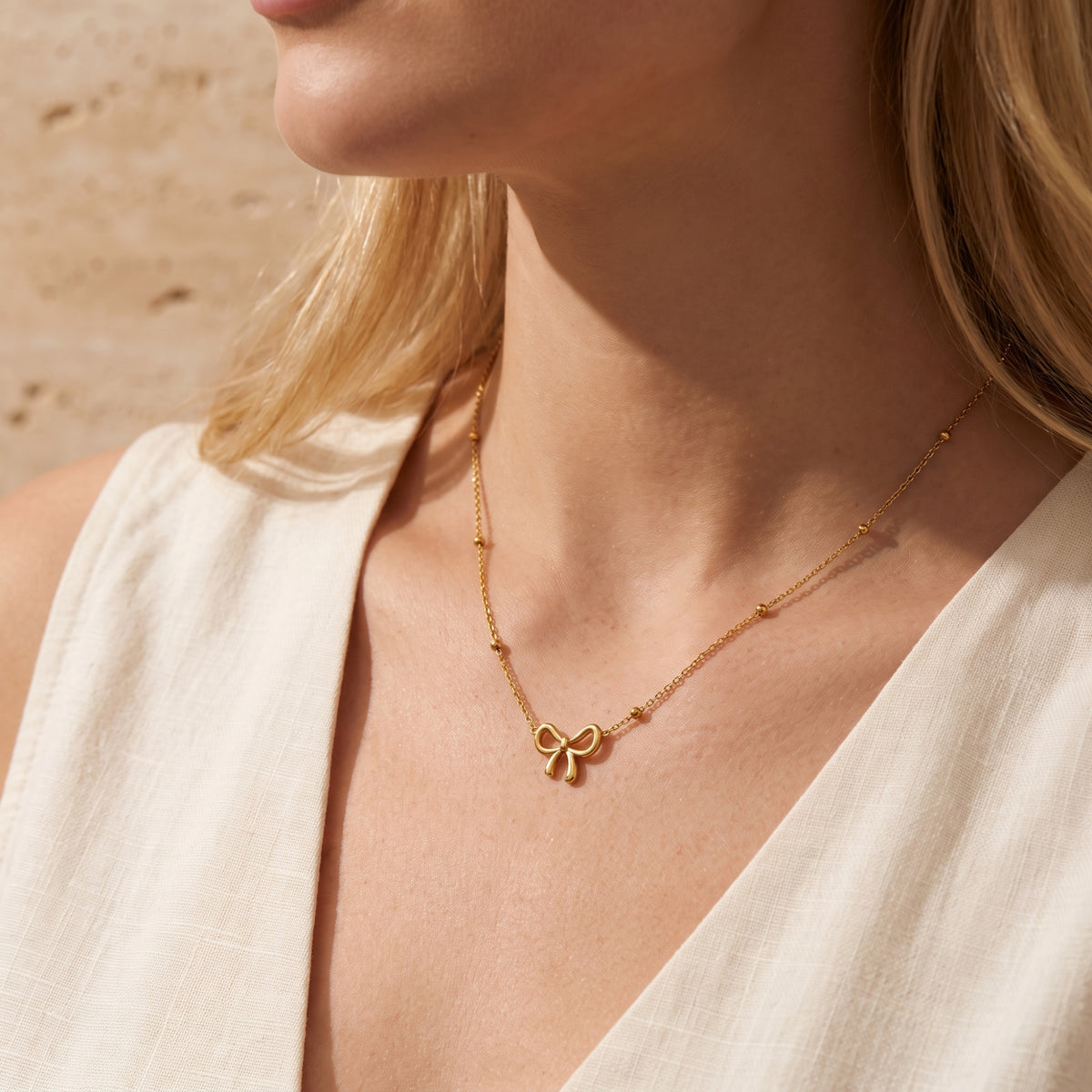 Model wearing the gold-tone bow station necklace, shown from the neck to chest in a light cream V-neck top.