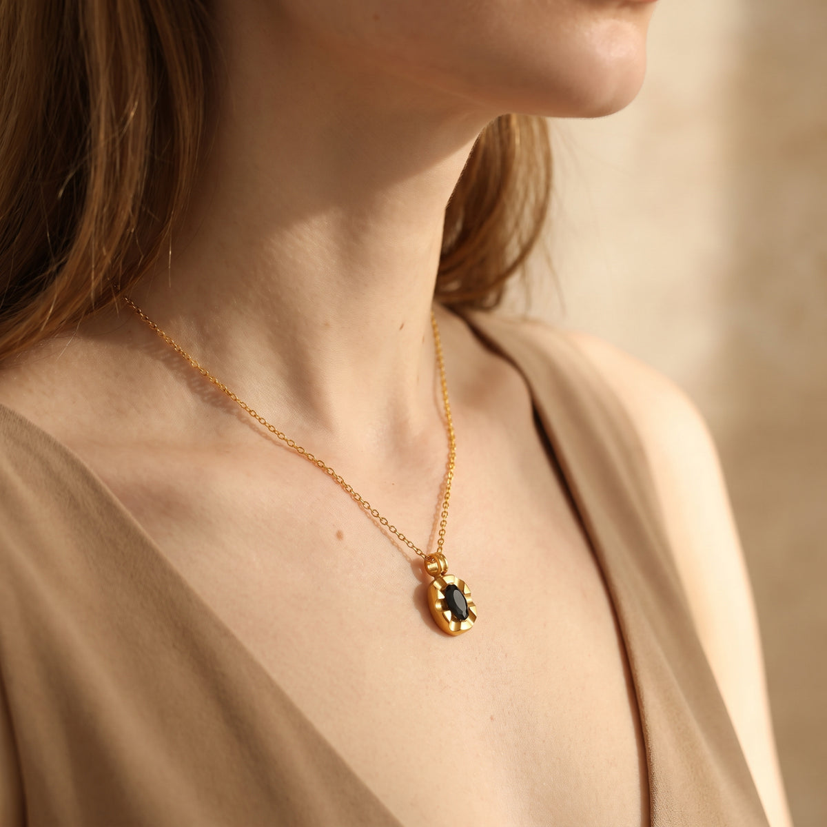 Side view of a model wearing the gold-tone chain and black oval medallion pendant over a beige V-neck garment.