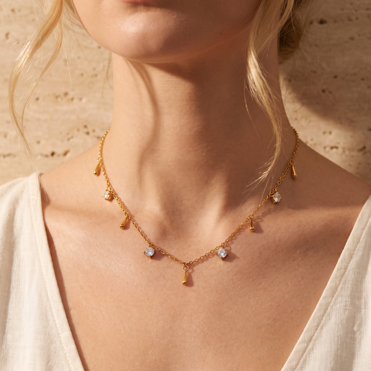 Front close-up of a woman wearing a gold-tone droplet necklace with clear stones and a cream V-neck top.