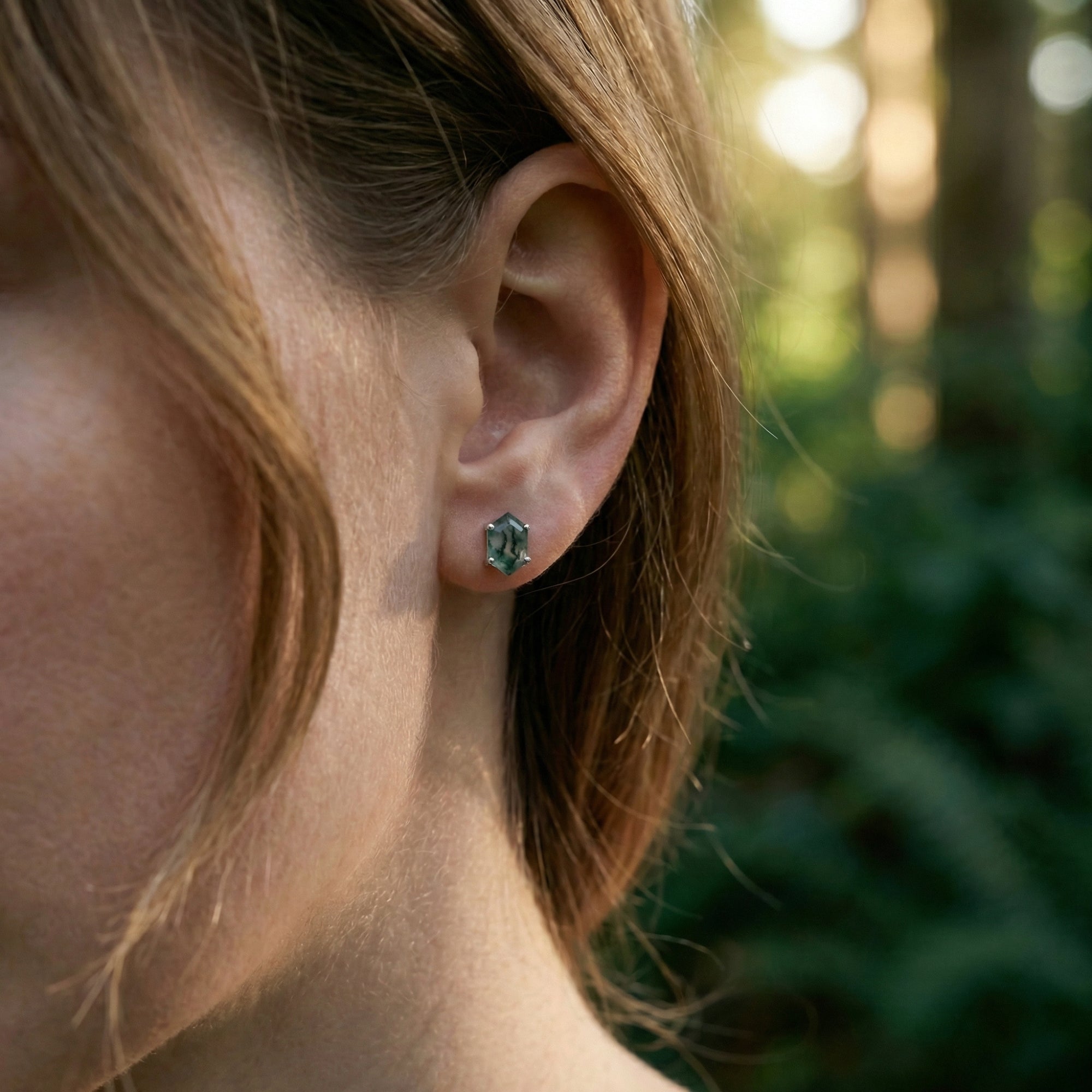 Profile view of a woman wearing a sterling silver hexagon moss agate stud earring in a natural forest setting.