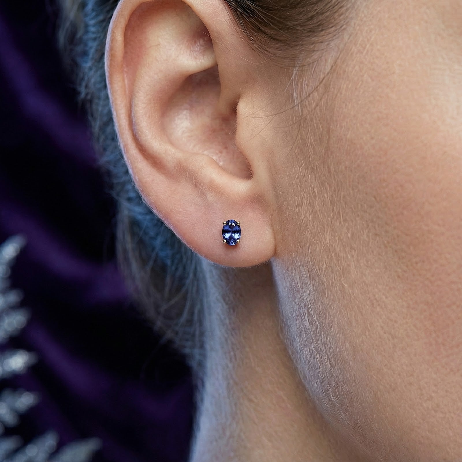 Close-up of a model wearing an 18K gold vermeil stud earring with an oval cut simulated tanzanite stone.
