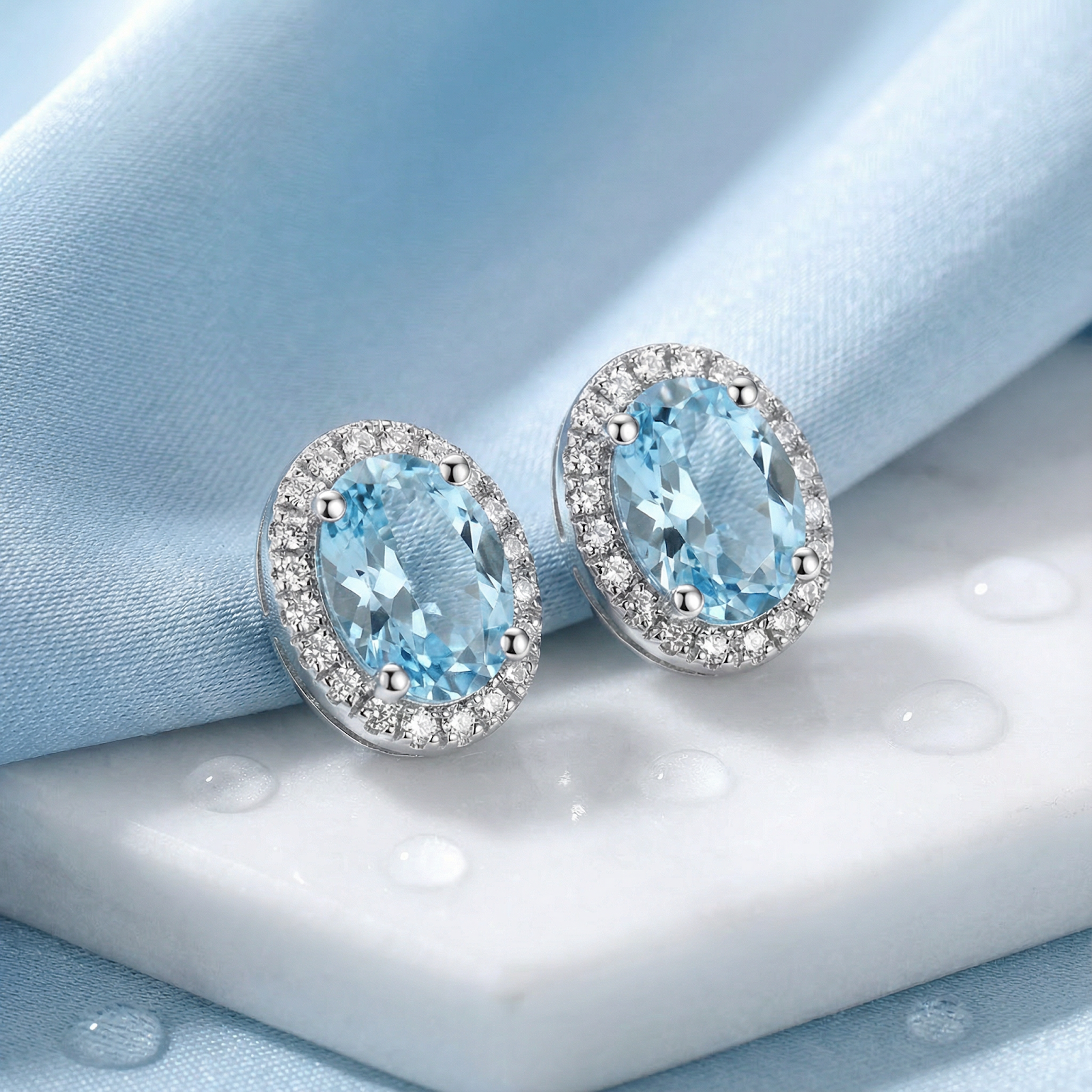 Oval simulated aquamarine halo stud earrings in sterling silver resting on a white marble and blue fabric background.