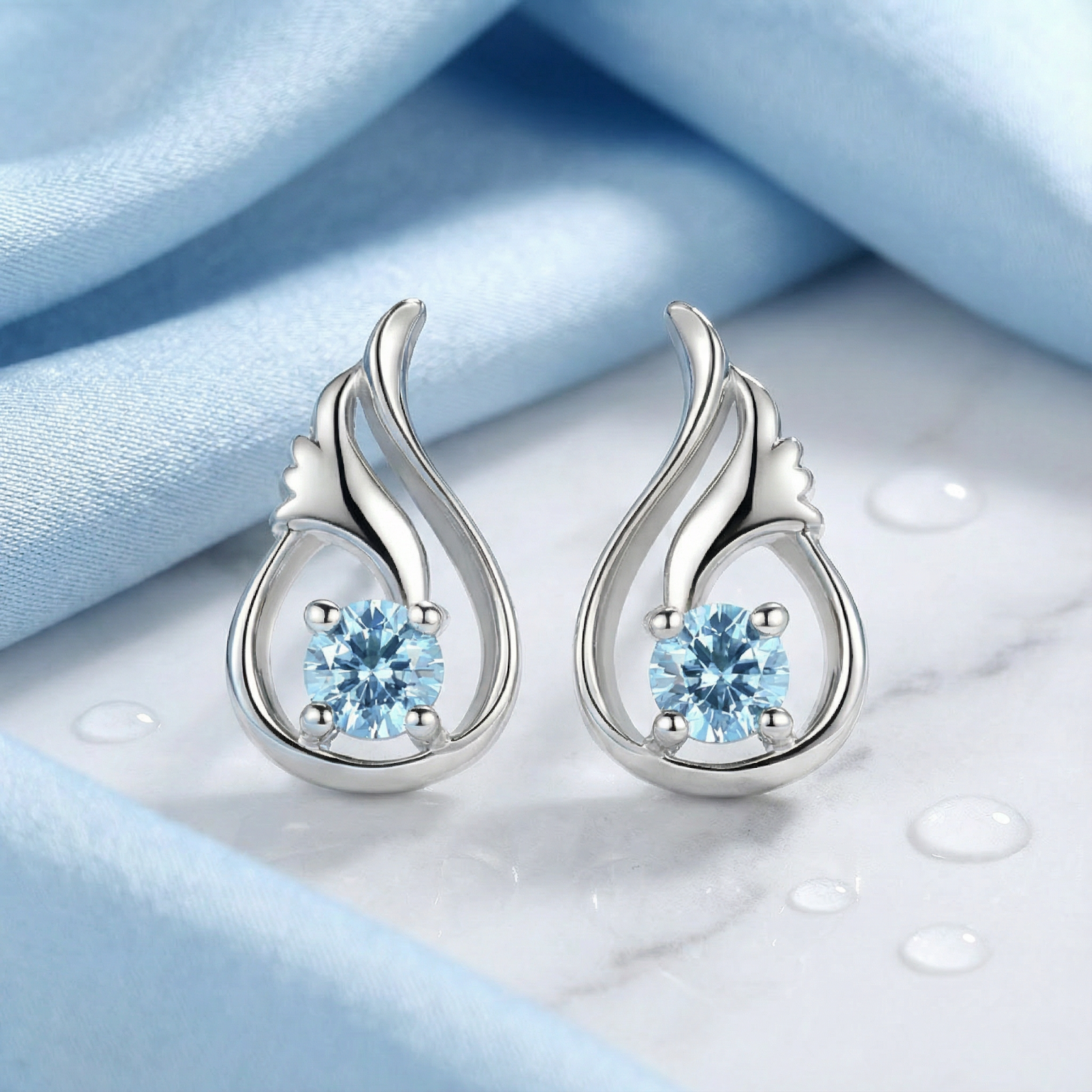 Front view of sterling silver drop earrings with round-cut light blue simulated aquamarine on blue fabric.