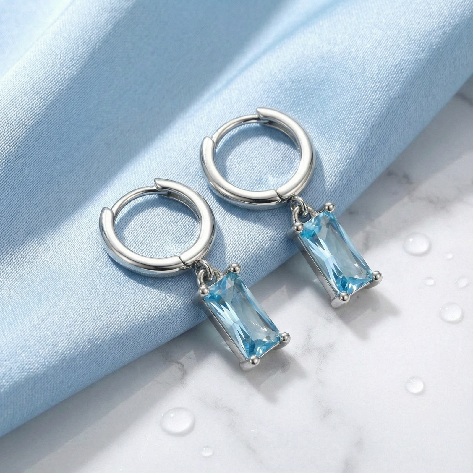 S925 sterling silver hoop earrings with rectangular-cut simulated aquamarine drops on blue fabric with water droplets.