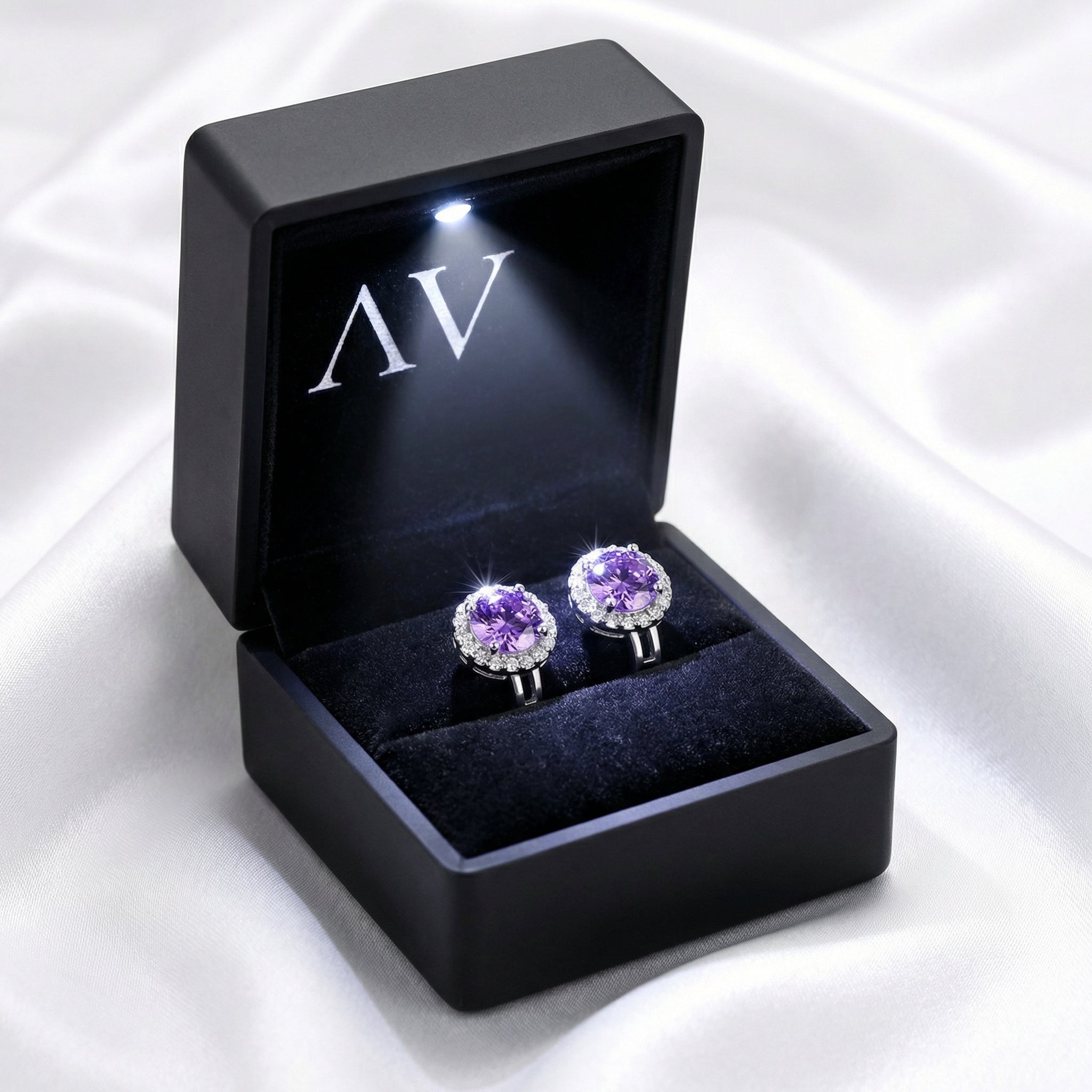 Round purple moissanite halo earrings in a black LED-lit gift box on white silk background.