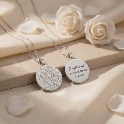Two silver-toned round pendants on cream fabric, one engraved with a star map and the other with custom text.