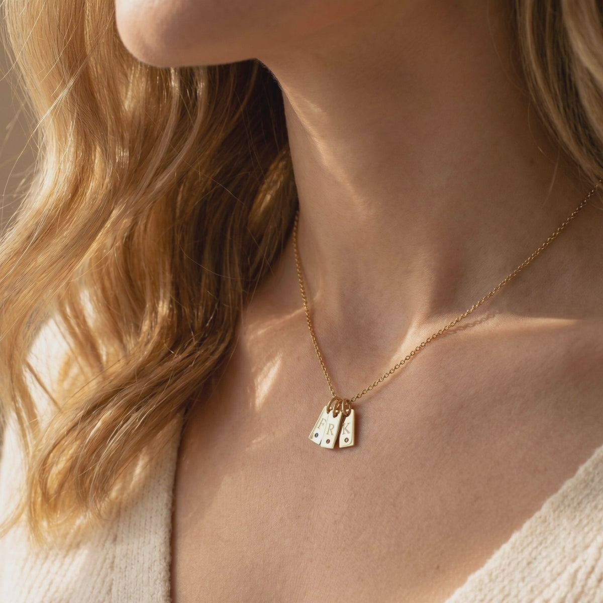 A woman wears a gold-tone necklace with three rectangular initial tags engraved F, R, and K, each with a small round stone.