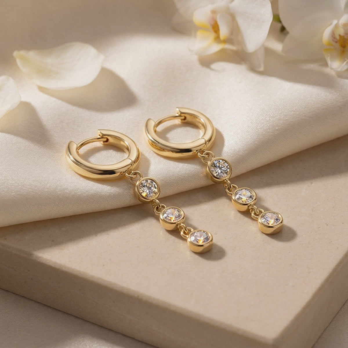 Pair of gold huggie hoop drop earrings with three round bezel-set stones each, displayed on a pale box beside white flowers.