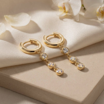 Pair of gold huggie hoop drop earrings with three round bezel-set stones each, displayed on a pale box beside white flowers.