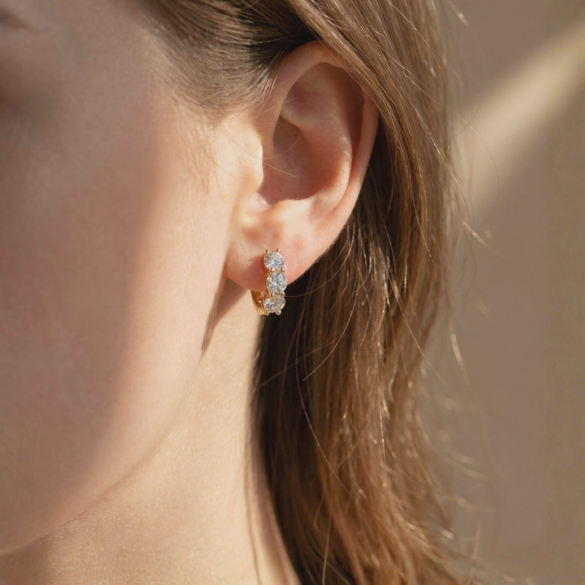Close-up of a person wearing one gold-toned huggie hoop earring with three round moissanite stones.