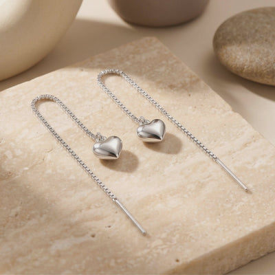 Pair of silver-toned box-chain heart threader earrings arranged on a textured beige stone surface.