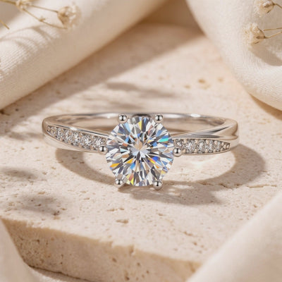 Close-up of a silver solitaire ring resting on a beige stone block, showing its six-prong setting and pavé band accents.