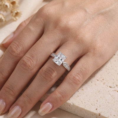 Silver ring with a radiant-cut clear center stone and embellished shoulders, shown worn on a finger.
