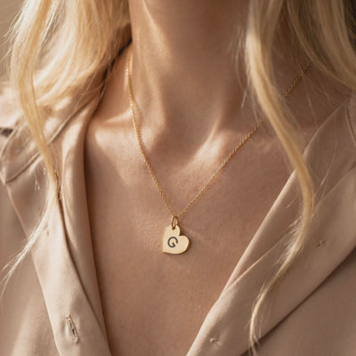 Gold-toned heart pendant engraved with the letter G, shown worn on a chain against a model's neck.