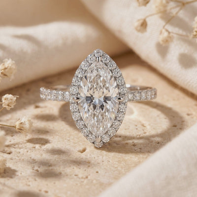 Close-up of a silver marquise moissanite halo ring with a pavé band displayed upright on a textured surface.