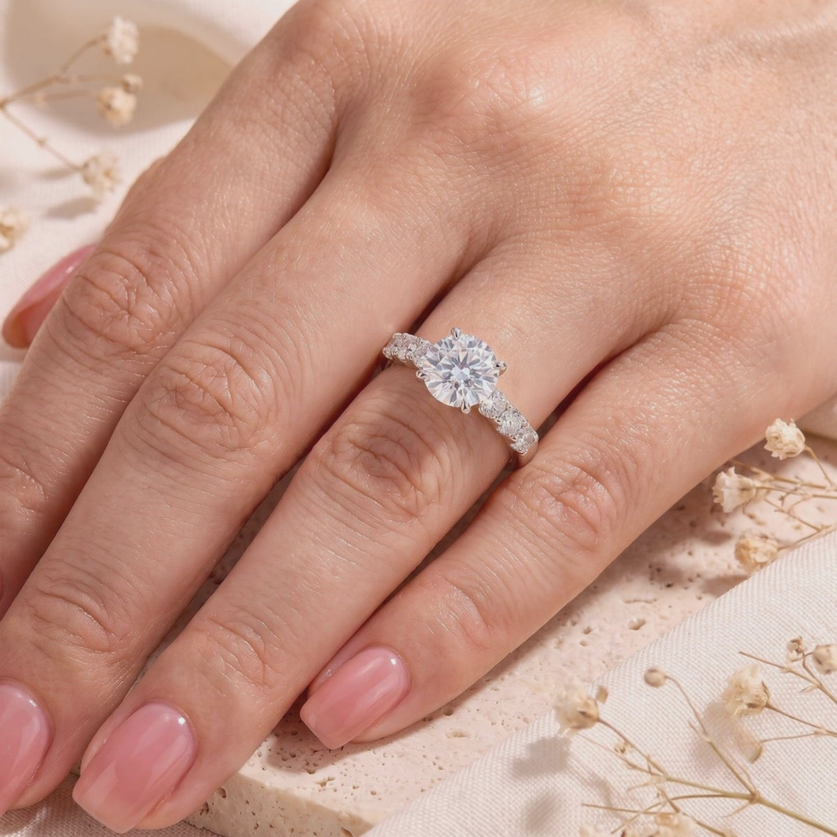 Silver ring with a round clear center stone and accent gems, displayed on a hand with glossy pink nails.