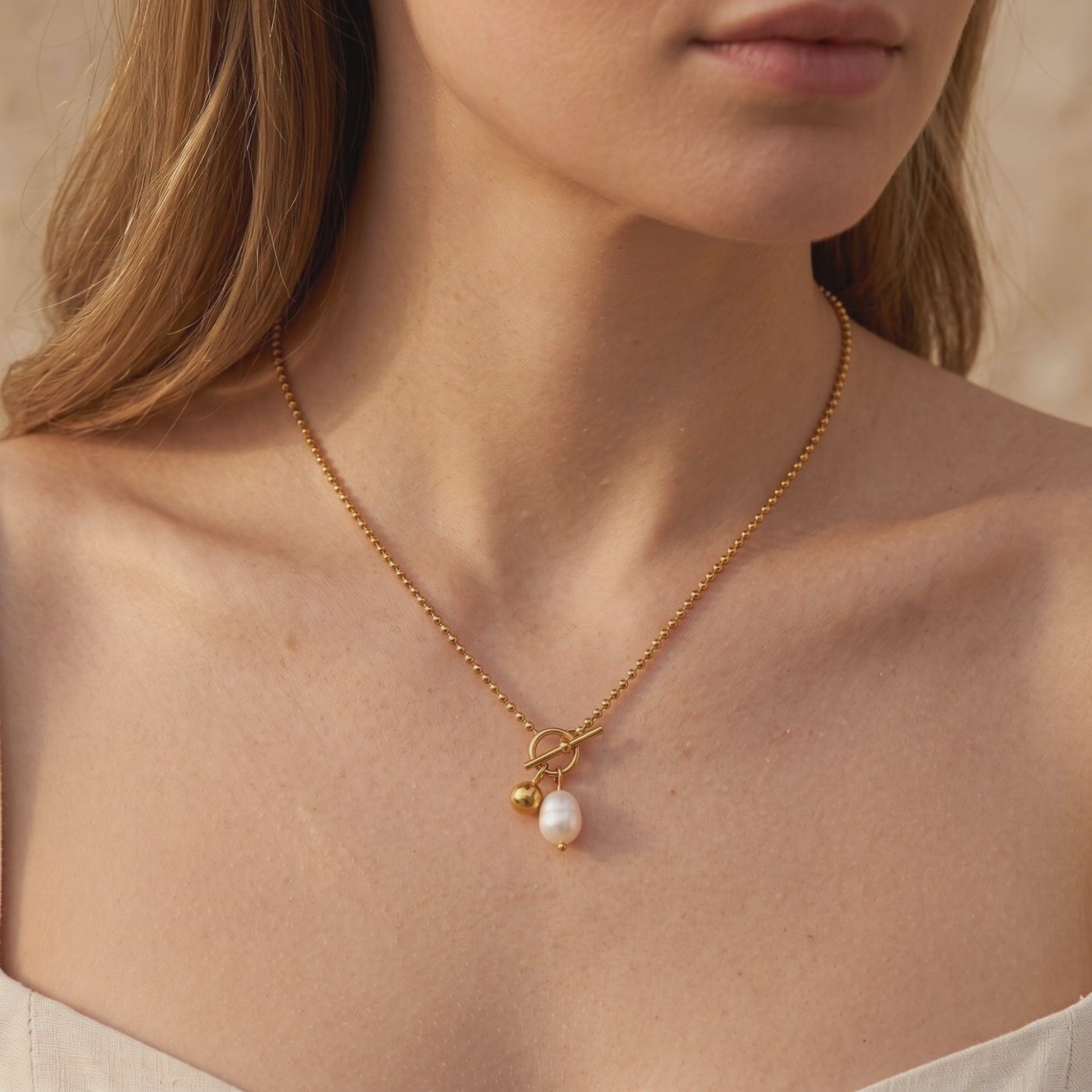 Gold-toned bead chain necklace worn on a model, with a front toggle clasp, pearl, and sphere charm.