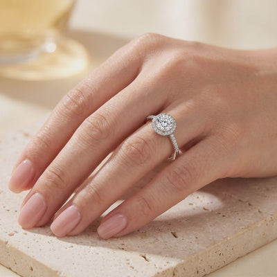 Round halo ring with a clear center stone and pavé band, shown worn on a hand.
