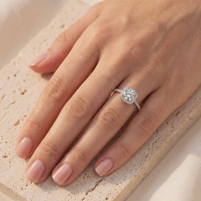 Silver halo ring with a round clear center stone, worn on a hand over a textured stone surface.