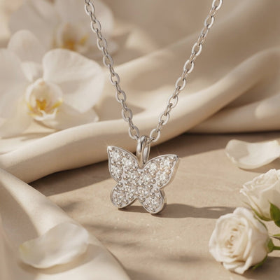 Close-up butterfly pendant with round clear stones on a chain, displayed on beige fabric with white flowers.