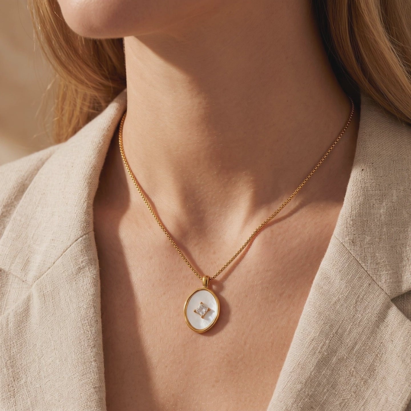 Gold-tone necklace with an oval mother-of-pearl pendant and square center stone, worn over a beige blazer.