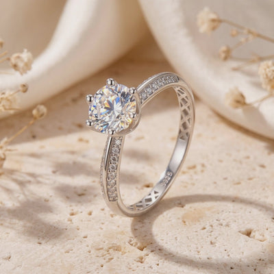 Close-up of a silver solitaire ring standing upright, showing its six-prong setting and pavé accents.