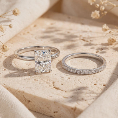 Silver-tone emerald-cut ring and matching pavé band displayed separately on a textured surface with dried flowers